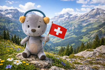Ein grauer Plüschbär mit Kopfhörern steht auf einem alpinen Berg in der Schweiz und hält eine Schweizer Flagge. Im Hintergrund sind blühende Bergwiesen, grüne Täler und schneebedeckte Alpen unter blauem Himmel zu sehen.