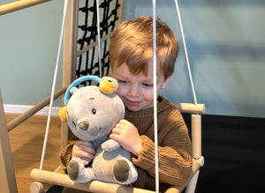 Ein kleines Kind sitzt auf einer Holzschaukel in einem Indoor-Spielbereich und hält Aaron, den grauen Bären mit blauem Kopfbügel und gelben Ohrwärmern, fest im Arm. Der Junge trägt einen braunen Strickpullover und lächelt verträumt, während er das Plüschtier an sich drückt. Der Hintergrund zeigt Kletterelemente aus Holz und Seilen.