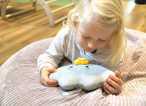 Ein Kleinkind liegt gemütlich auf einem weichen Kissen und hält Lenabie Lina (Wolke) sanft mit beiden Händen; sie schaut Lina neugierig an und lauscht dabei einer Hörspielwiedergabe von Lina, welche über die Lenabo App bequem von Eltern eingestellt und gesteuert wird. Das Kind blickt ruhig und aufmerksam auf Lina herab, wodurch eine warme, beruhigende Stimmung entsteht.